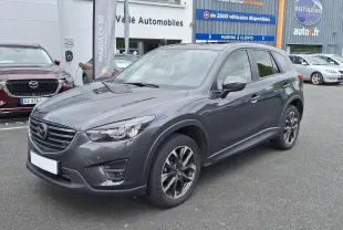 Vue 3/4 avant droit d'un Mazda CX-5 gris métallisé garé devant un concessionnaire avec jantes aluminium noires.