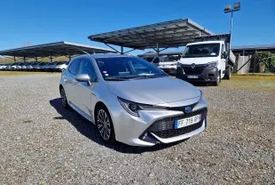 Toyota Corolla hybride gris clair métal vue 3/4 avant droit sur parking extérieur avec plusieurs véhicules en arrière-plan.