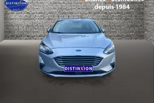 Vue frontale d'une Ford Focus SW gris clair avec calandre chromée et phares allumés sur fond de mur en briques.