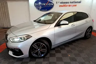 BMW Série 1 gris clair vue 3/4 avant droit, phares LED allumés et jantes sport noires en showroom