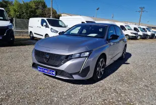 Peugeot 308 gris Artense vue 3/4 avant droit sur parking gravillonné avec plusieurs fourgons blancs en arrière-plan.