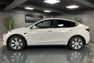 Profil côté gauche d'une Tesla Model Y blanche nacrée RWD 2022 dans un showroom avec mur gris et plantes.