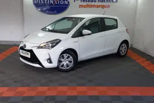 Toyota Yaris blanche vue de profil côté gauche en intérieur, avec phares allumés et logo hybride visible sur l'aile avant.