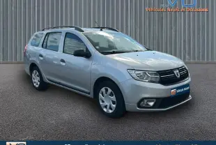 Vue 3/4 avant droite d'une Dacia Logan MCV gris clair avec barres de toit et jantes 15 pouces, stationnée sur sol bitumé.