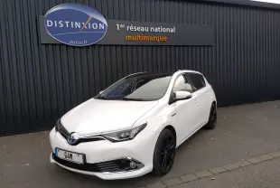 Toyota Auris hybride blanche vue en 3/4 avant droit, avec jantes noires et calandre chromée devant un mur noir.