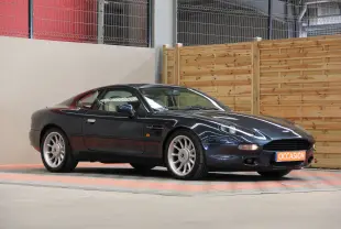 Aston Martin DB7 bleu marine vue 3/4 avant droit, avec jantes argentées et plaque "OCCASION" orange visible.