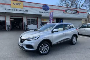 Renault Kadjar Blue dCi 115 Business gris clair en 3/4 avant droit devant un garage avec panneaux publicitaires.
