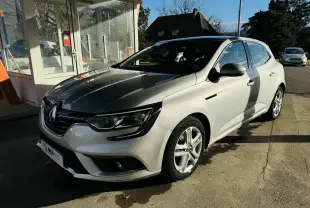 Renault Mégane IV berline gris vue 3/4 avant droit, stationnée devant un bâtiment vitré par temps ensoleillé.