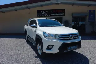 Toyota Hilux 4x4 blanc en 3/4 avant droit, double cabine, avec calandre chromée et barres de toit visibles.