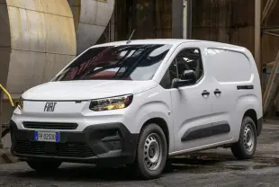 Fourgon Fiat Doblo blanc en 3/4 avant droit, avec calandre noire et feux allumés dans un environnement industriel.