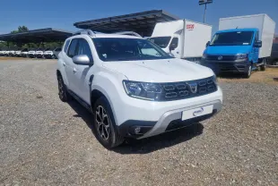 Dacia Duster blanc en 3/4 avant droit, avec calandre noire et jantes alliage, stationné sur un sol gravillonné.