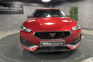 Vue frontale d'une CUPRA Leon Sportstourer rouge Désir avec calandre noire et logo cuivré dans un showroom.