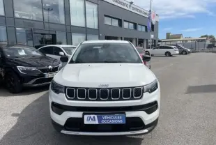 Vue frontale d’un Jeep Compass blanc 2024 avec calandre à sept fentes et plaque « Véhicules Occasions » devant un garage.