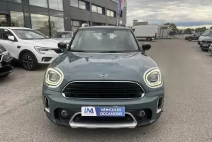 Vue frontale d'une MINI Countryman Cooper 136 gris avec phares allumés sur un parking de véhicules d'occasion.