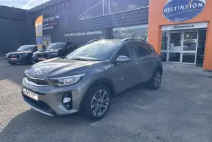 KIA Stonic brun Sienne en vue 3/4 avant droit, avec jantes alliage et barres de toit visibles, devant un bâtiment commercial.