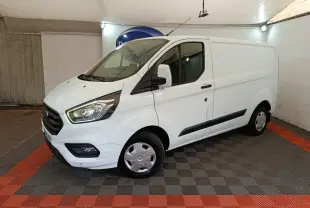 Vue 3/4 avant droit d'un fourgon Ford Transit Custom blanc avec calandre noire et feux avant allumés.